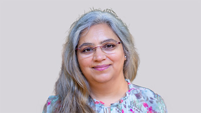 Jyoti Taneja, a person with long gray hair wearing glasses and a blue, pink, and purple top, against a gray background.