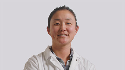 Michelle Lu, a person with dark hair pulled back wearing a white coat over a gray collared shirt, against a gray background.