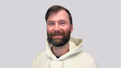 Nathan Good, a person with short dark hair and beard wearing a yellow hoodie against a gray background.