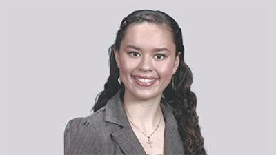 Patricia McNeil, a person with long dark wavy hair wearing a gray blazer and necklace, against a gray background.