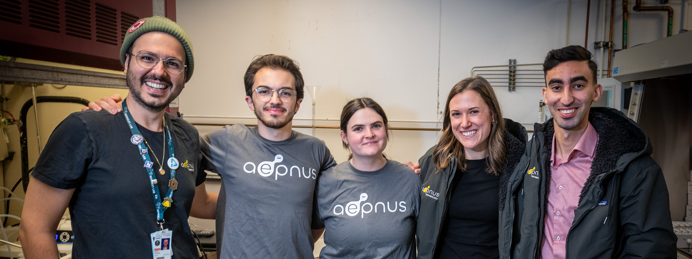 A diverse group of fellows stand together, arm in arm, and smile.