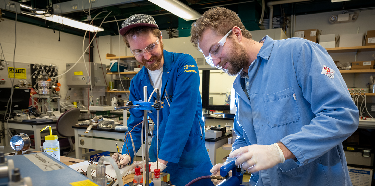 Two men in lab coats with safety goggles work on an experimental setup in a workshop.