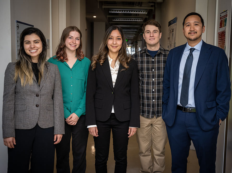 A group of five young people in smart business clothes stand in a hallway smiling.