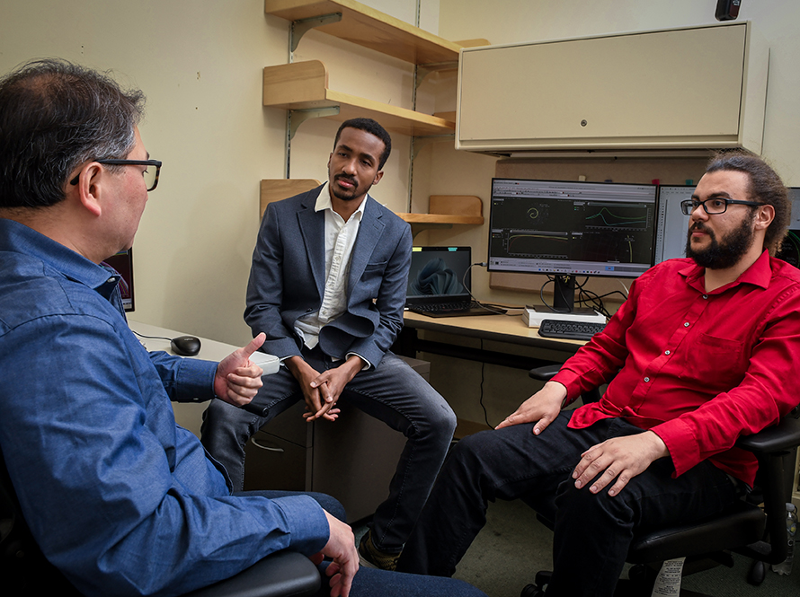 A diverse group of men sit in an office talking to each other.