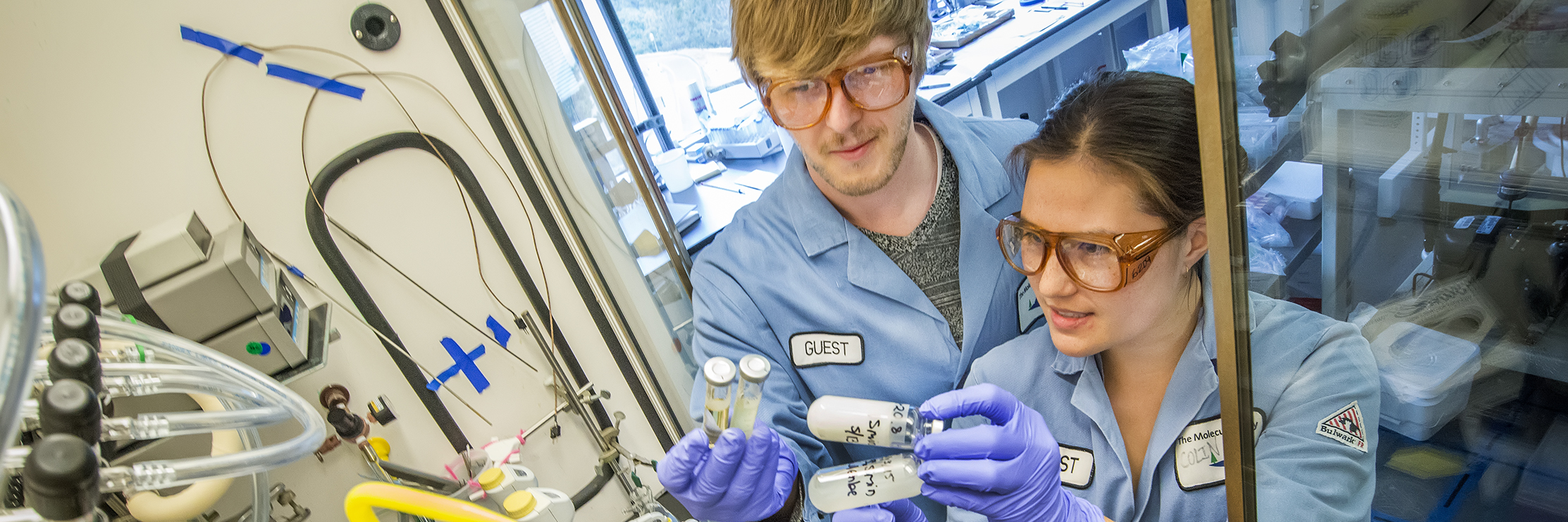 A man and woman in lab coats work in a laboratory, holding vials to install them in a fume hood.