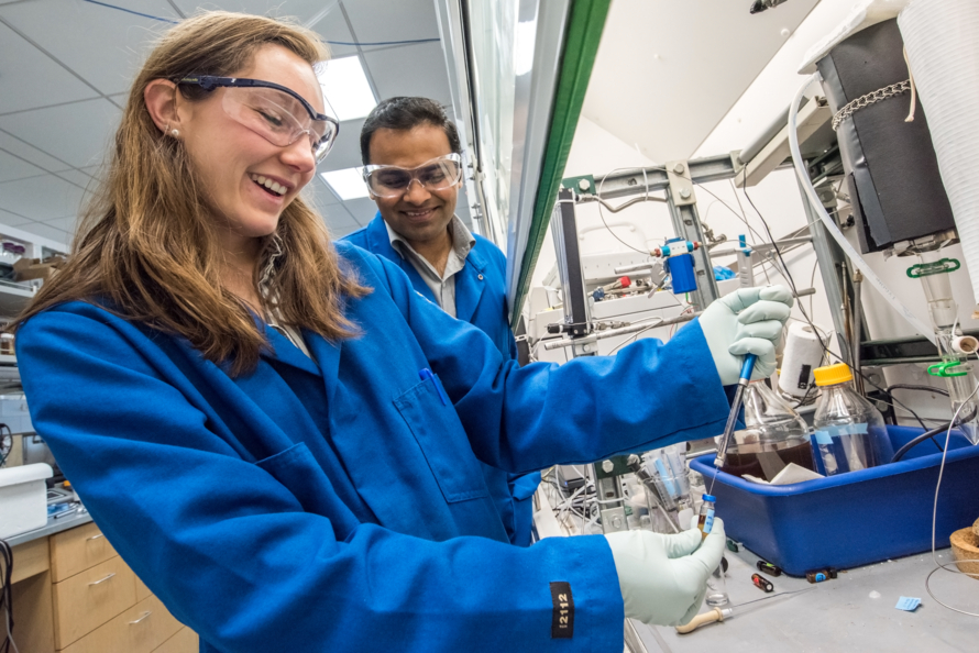 A woman and a man in lab coats and safety glasses fill vials in a laboratory.